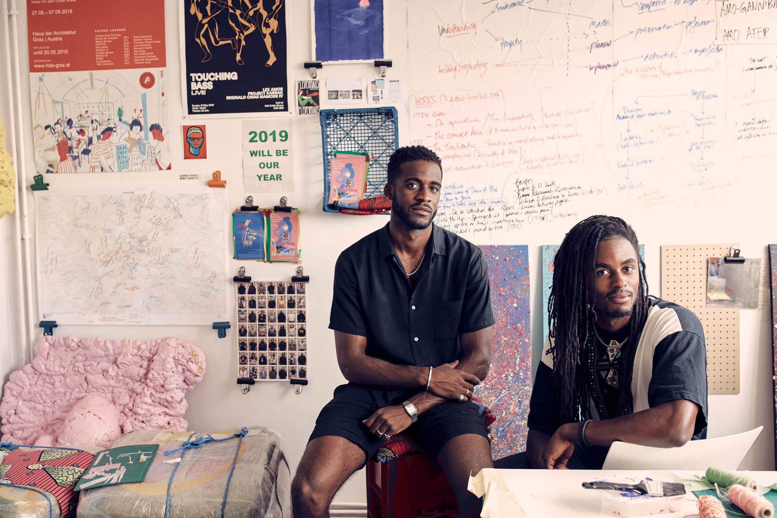 Posed headshot portrait of two men sat down in an art studio, looking straight at the camera with slight smiles