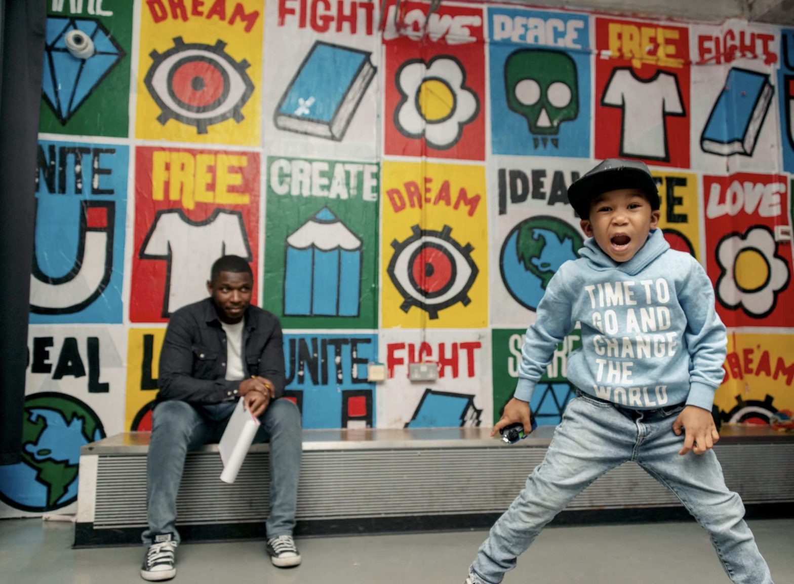 Image of Yomi Sode with his son Noah in the foreground. The background is a wall with colourful illustrated posters titled "dream", "free", "love", "fight", "peace", "creat", "unite"